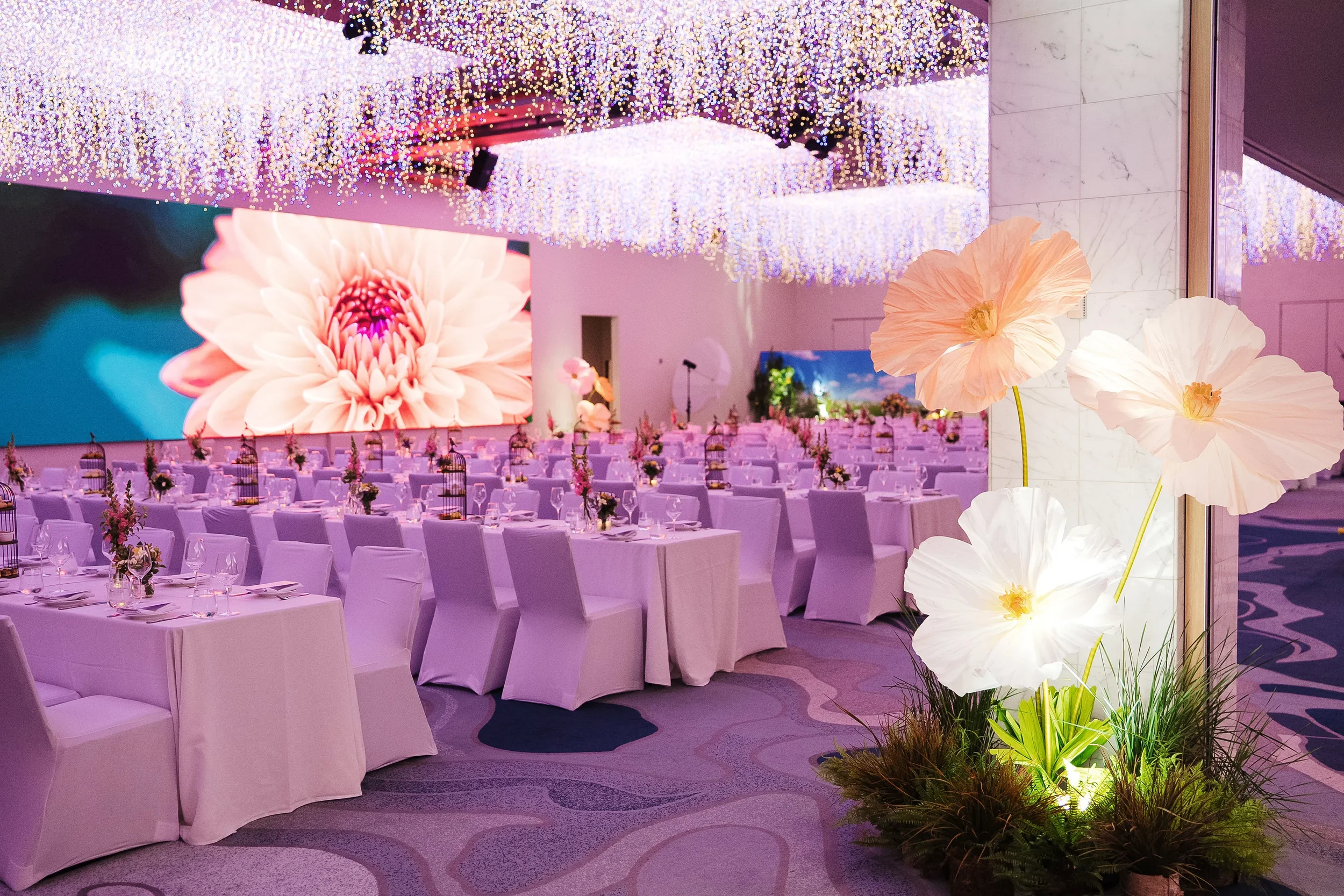 Elegant banquet hall decorated with pink and white flowers, hanging crystal chandeliers, and large LED screen displaying a close-up of a flower.