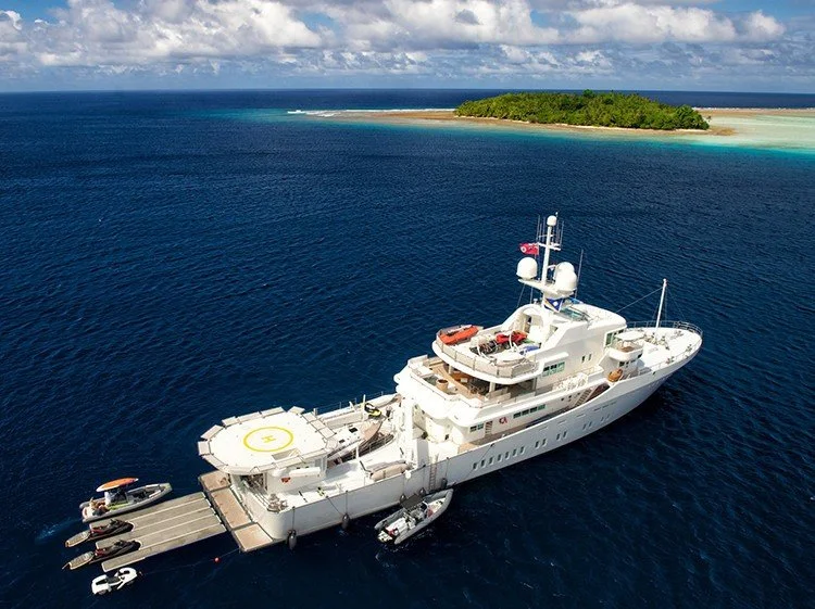 James Russ lighting. A large white yacht anchored in deep blue ocean near a tropical island with sandy beaches and lush green trees.