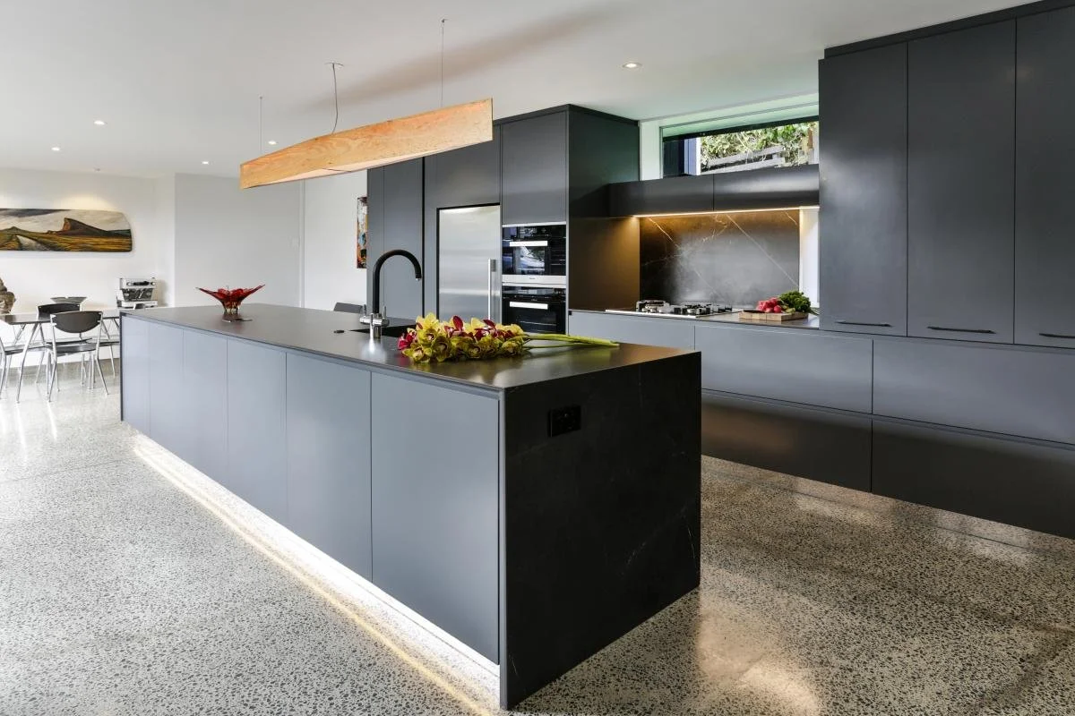 Modern kitchen with black and gray cabinetry, an island with a black countertop, built-in appliances, and a black vent hood over the stove. There is a window above the counter, and decorative items like flowers and a red bowl on the island.