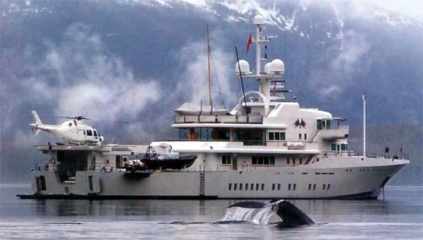 James Russ lighting. Large yacht with a helicopter on the deck and a whale surfacing nearby in the water, mountains in the background.