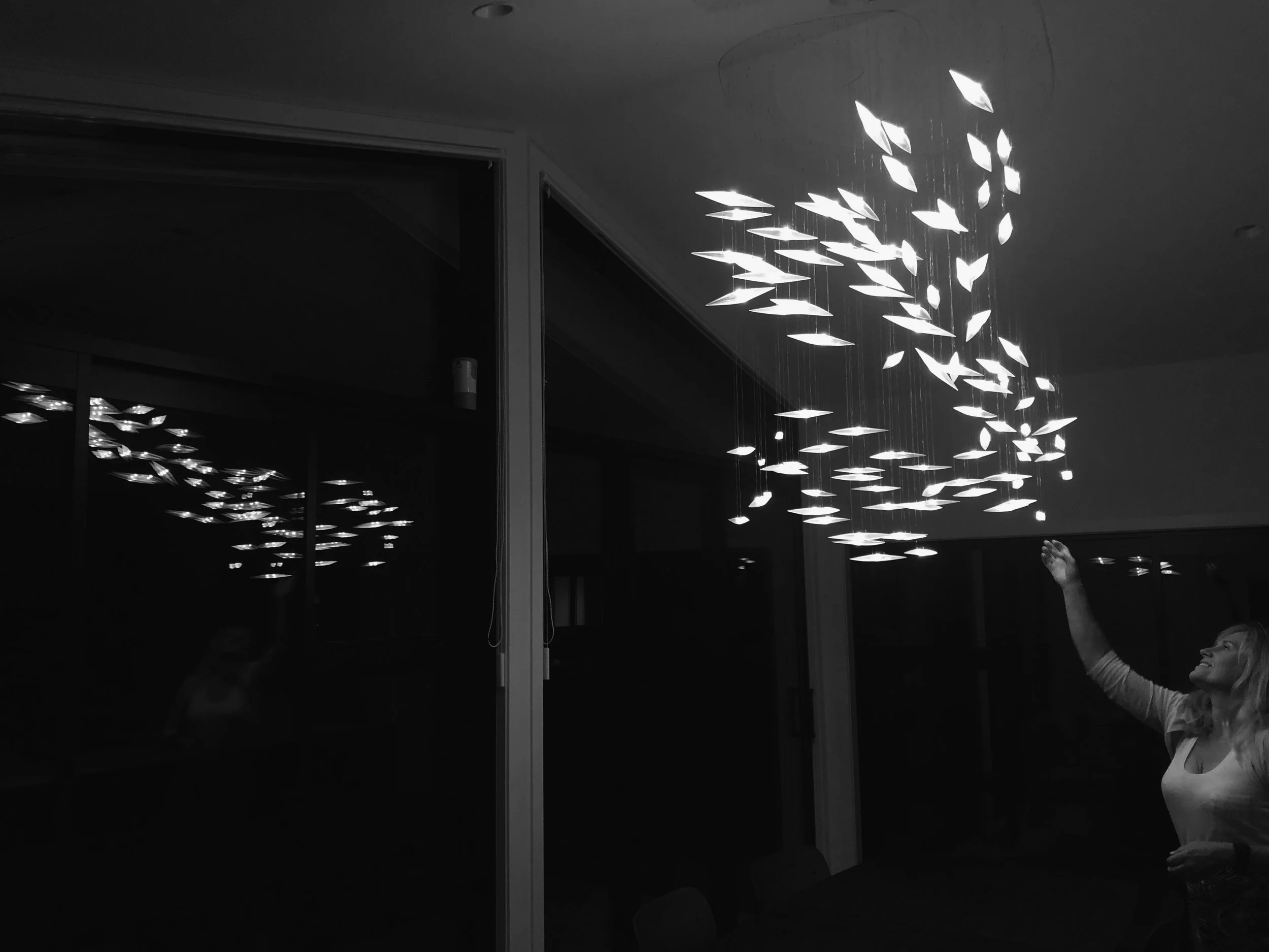 A woman reaches up to touch or interact with an illuminated, artistic ceiling light fixture in a dark room. The light fixture consists of multiple elongated, leaf-shaped lights hanging at varying lengths.