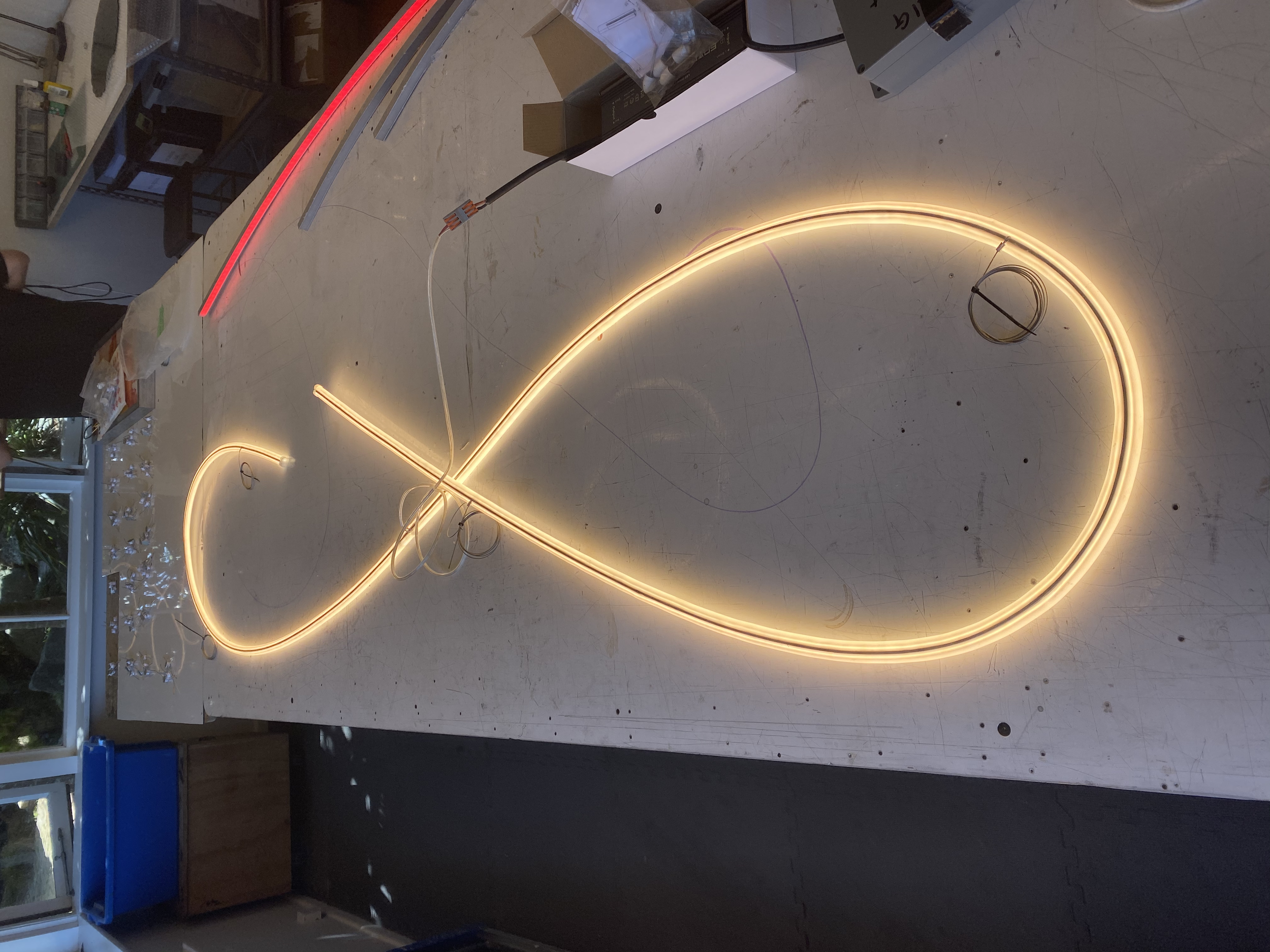 James Russ Sensory Sculpture. Illuminated neon light in a flowing, cursive design on a workbench, with electrical wires and tools nearby.