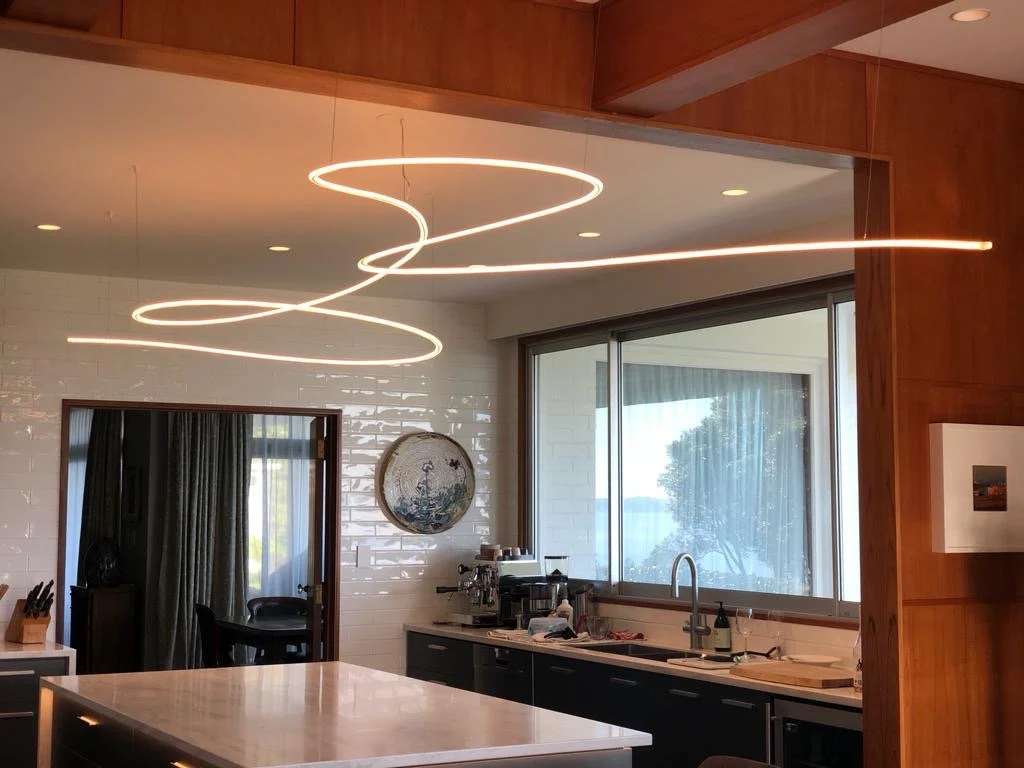 James Russ Sensory Sculpture. Modern kitchen with pendant light fixture hanging over the island, white tile backsplash, large window showing outdoor view, and dark cabinetry.