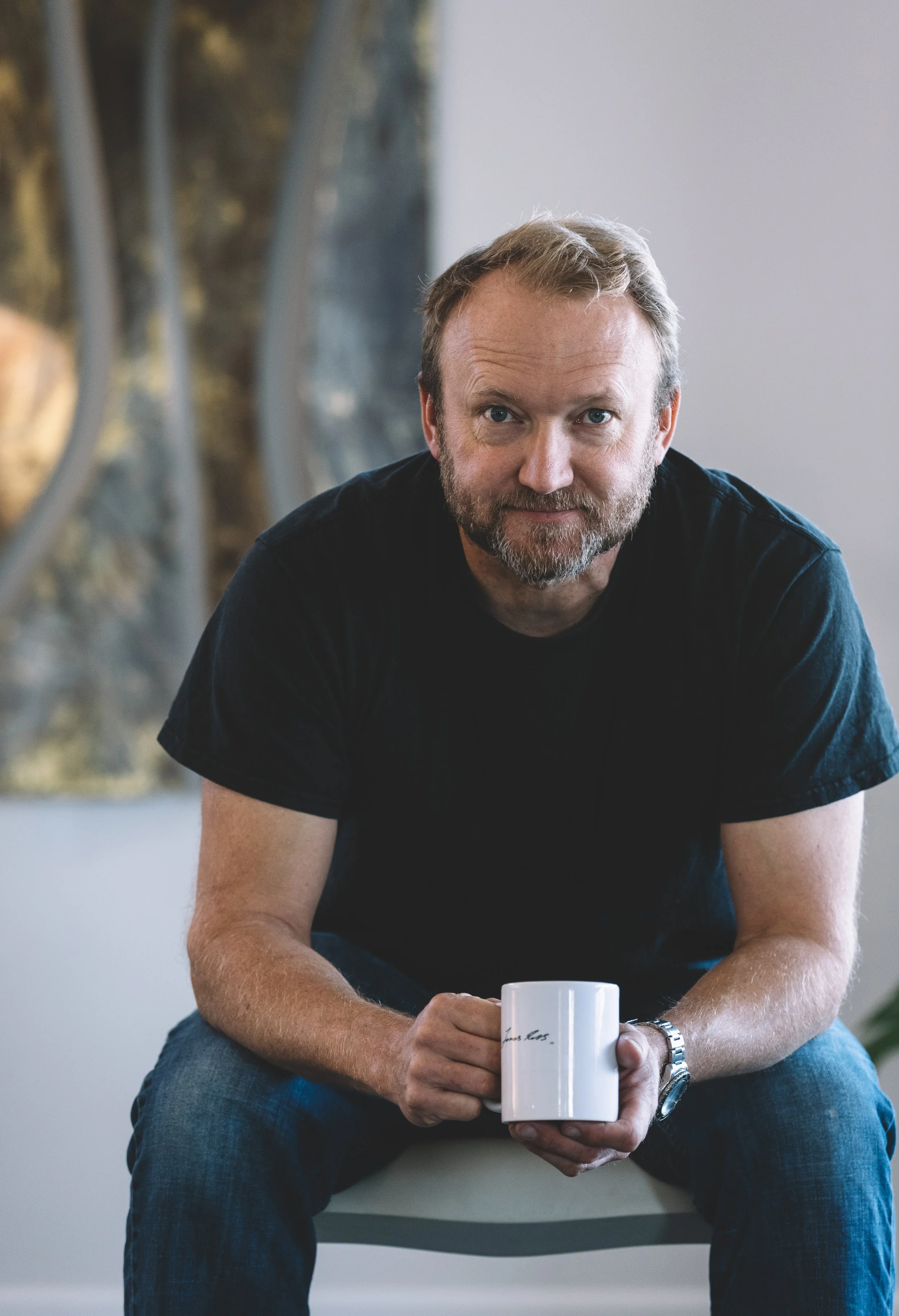 A man with a beard and mustache wearing a black t-shirt sitting on a chair, holding a white mug, with abstract art in the background.