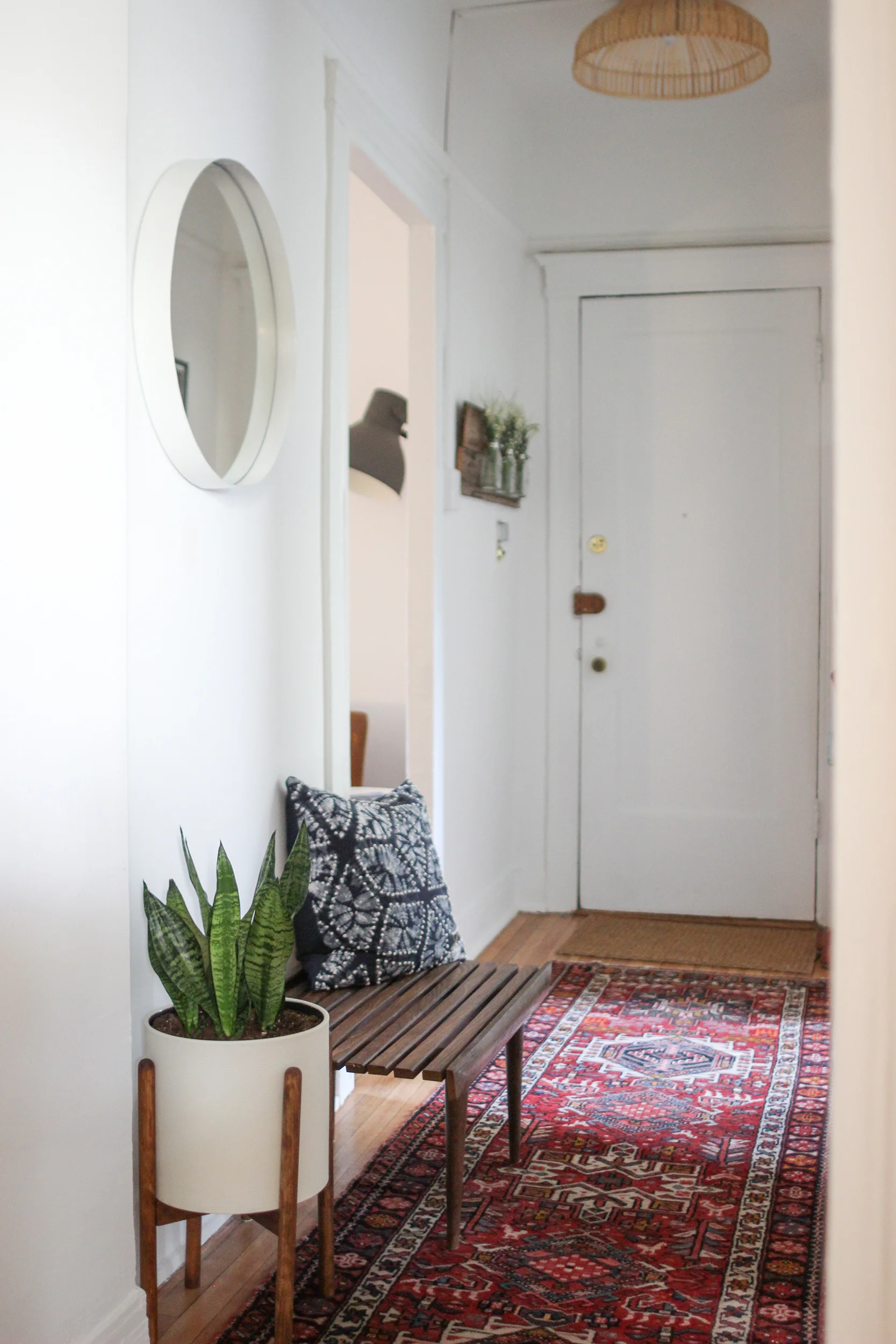  Hallway of an apartment with a colorful rug, plant, wooden bench, mirror, and door 