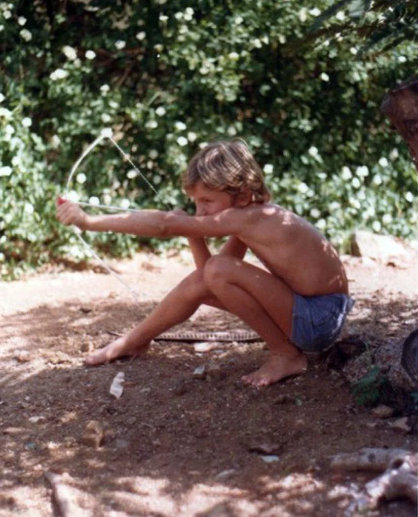 Young boy pulling bow and arrow on warm sand.