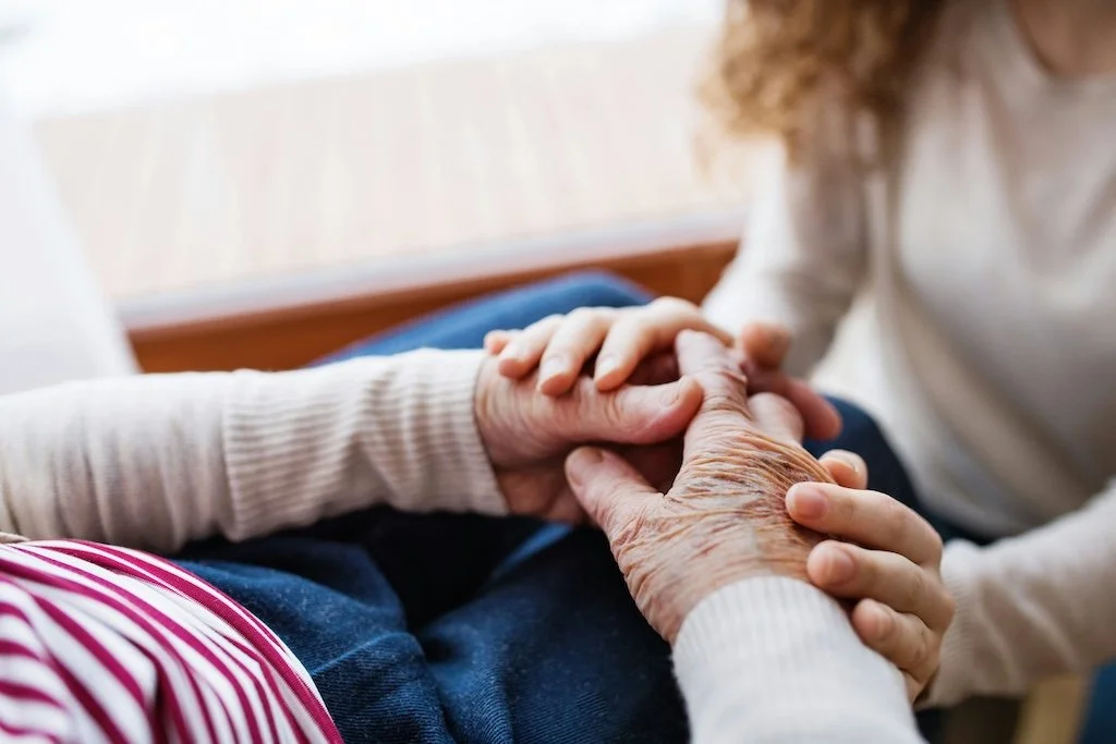 Elderly hands folded in lap, being gently held by young hands