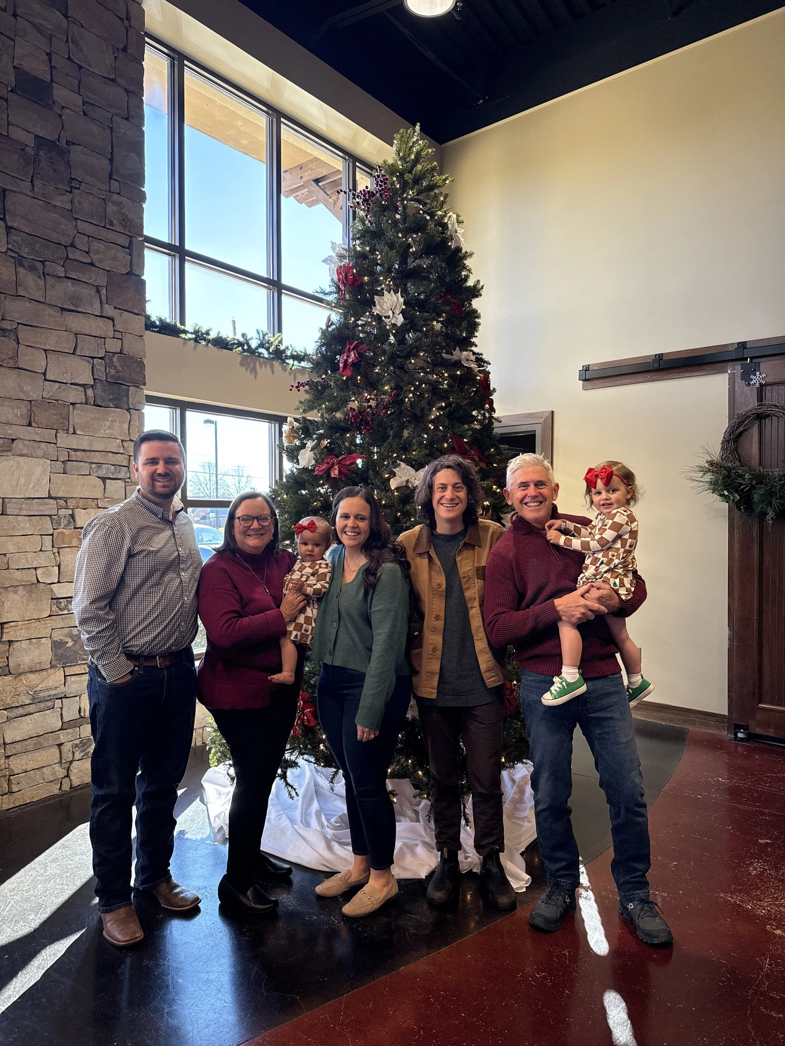 A group of seven people, including two young children, stands in front of a decorated Christmas tree inside a building with large windows and stone and wood wall accents. They are smiling and dressed in casual clothing.