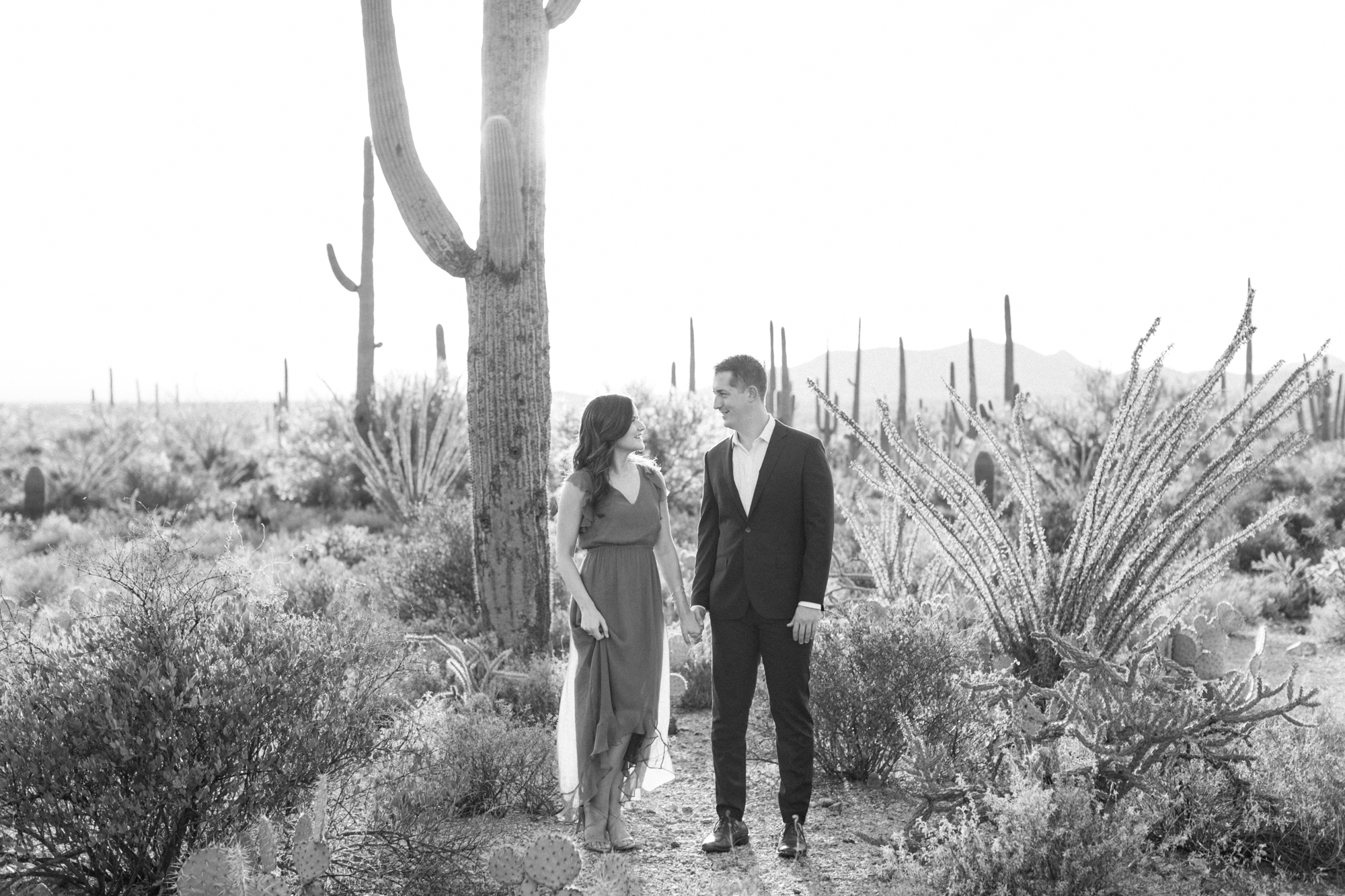 Black and white photo of a couple holding hands, looking at each other at their desert engagement session. 