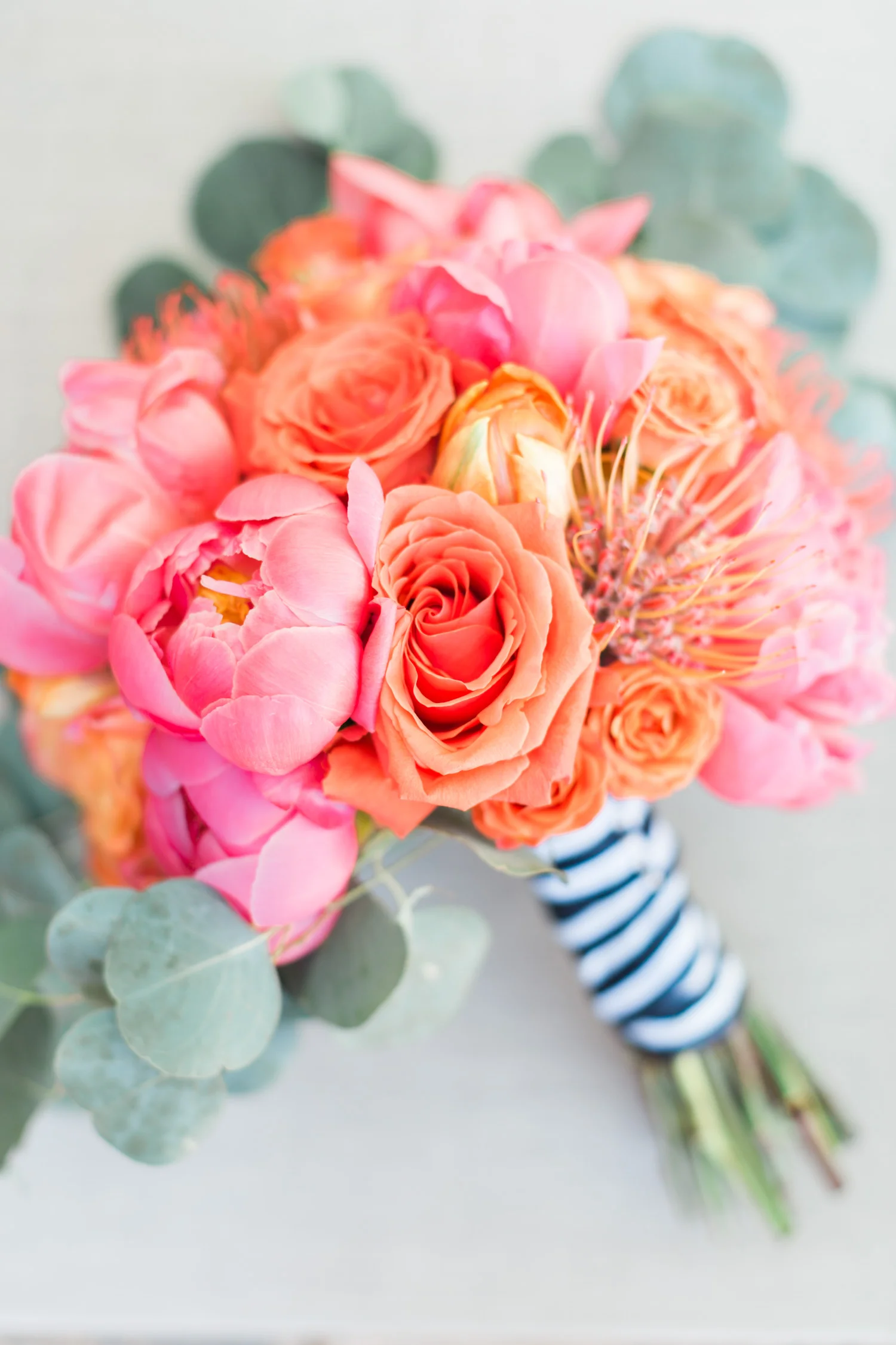 Beautiful bridal bouquet with pink peonies, protea pincushion, salmon and orange roses tied with a striped navy and white ribbon