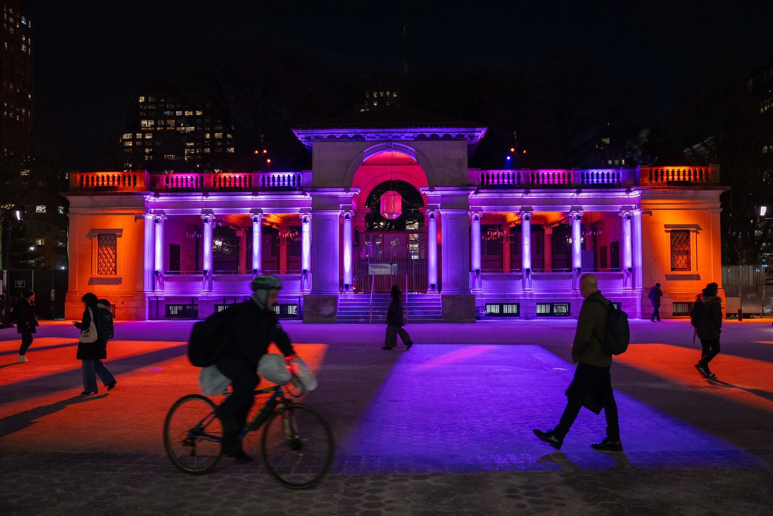 Outdoor architectural lighting traced onto a 3 story pavilion with symmetrical columns on each side, center opening with a reflective pendant. Silhouettes of people walking, standing, and a person on a bicycle.