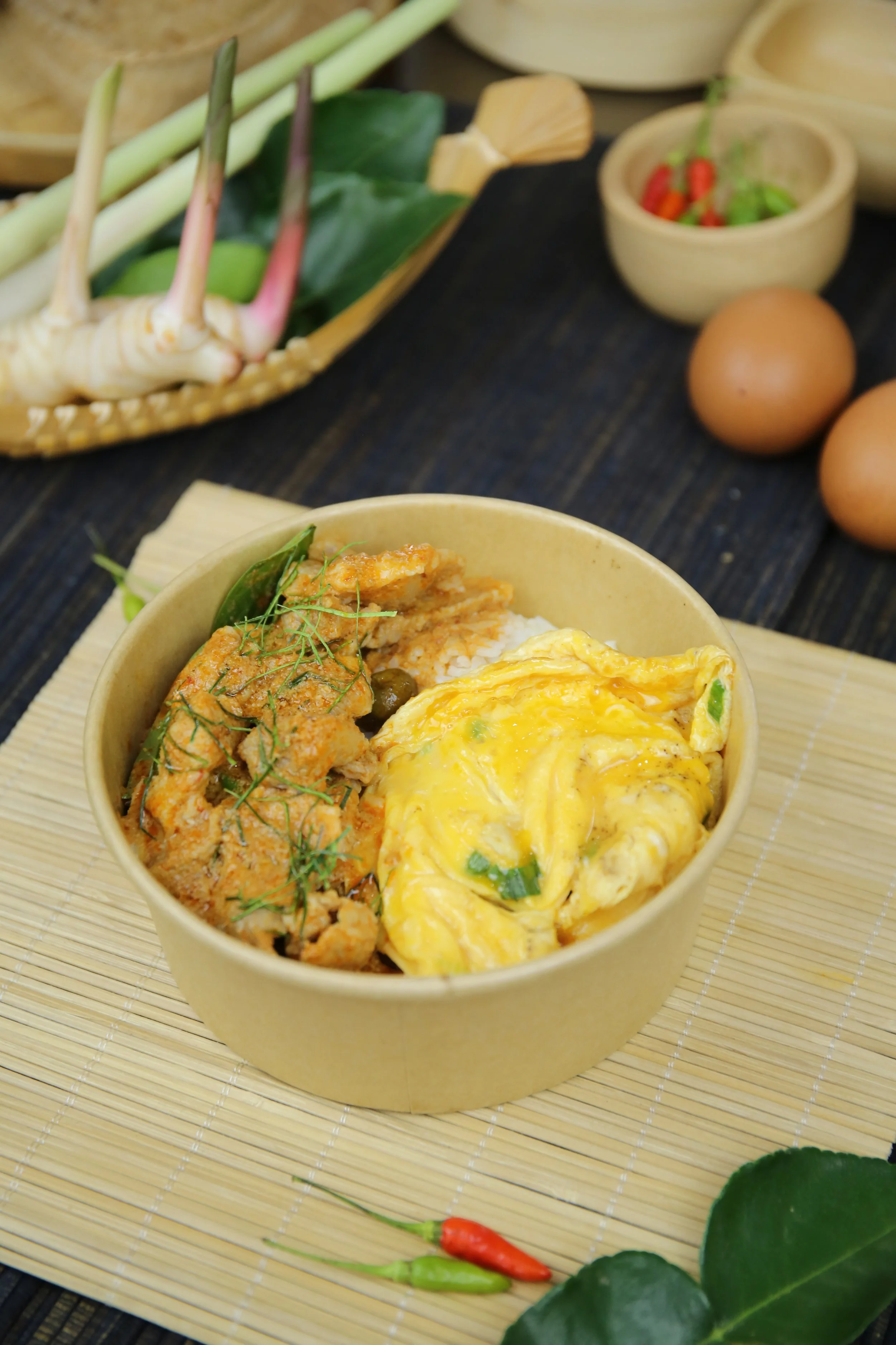 Bowl of rice with fried chicken, scrambled eggs, and vegetables, placed on a bamboo mat, with some eggs, a small bowl of herbs, and chili peppers in the background on a dark wooden surface.