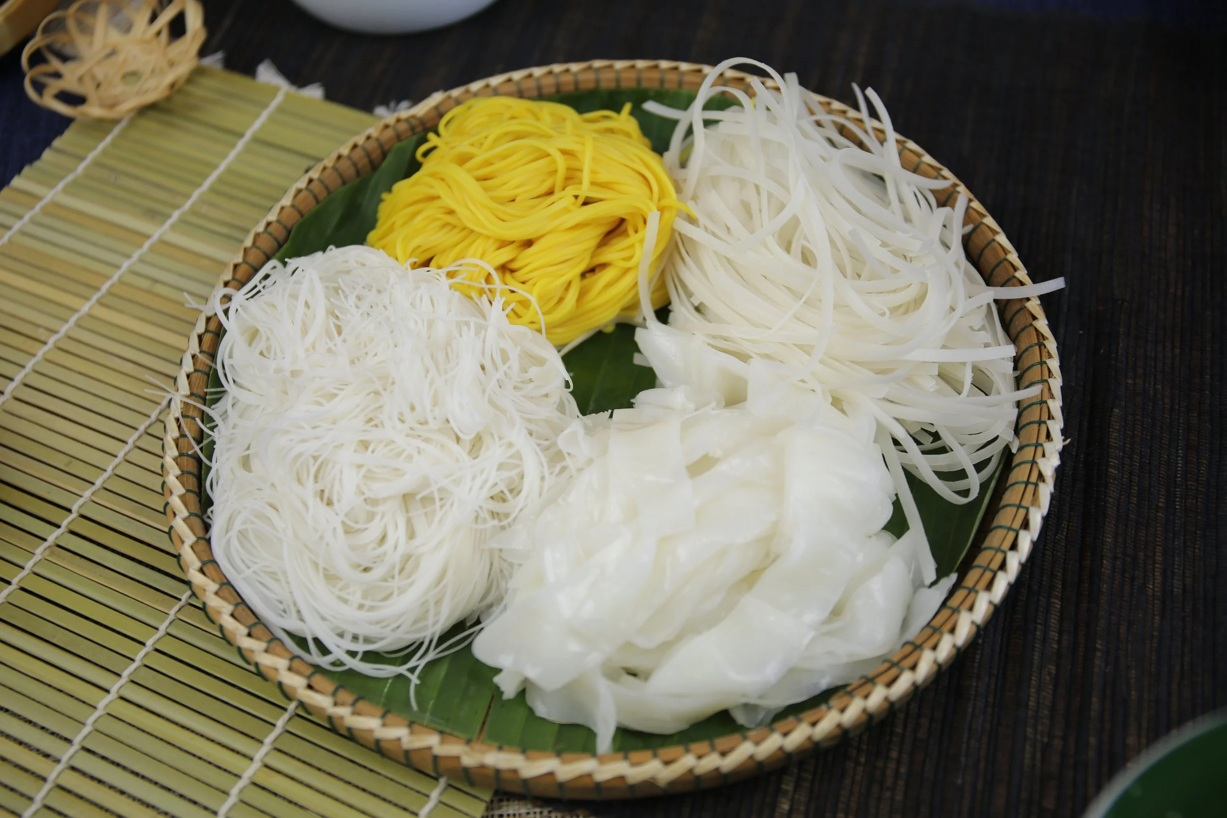 Basket with bundles of uncooked rice noodles in white and yellow, on a green banana leaf, on a bamboo mat.
