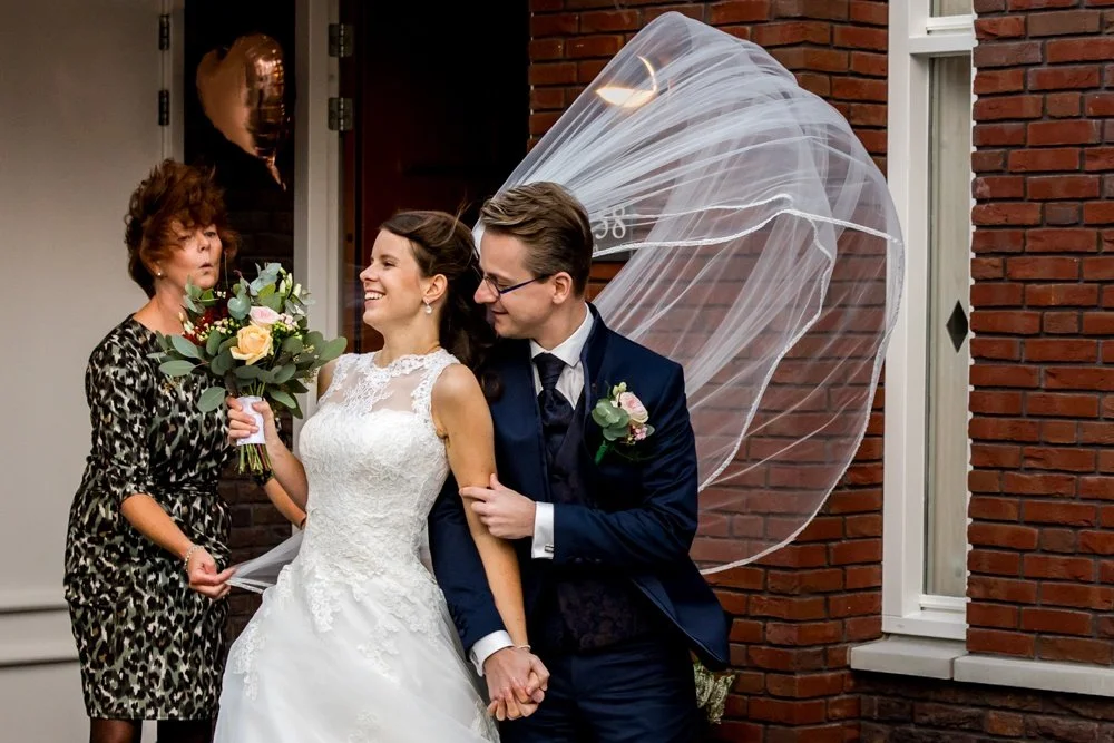 voorbeeld van journalistieke trouwfotografie: bruid komt bij de first look naar buiten en de sluier vangt wind en waait weg wat voor een humoristische situatie zorgt samen met de moeder van de bruid en bruidegom in Leiden