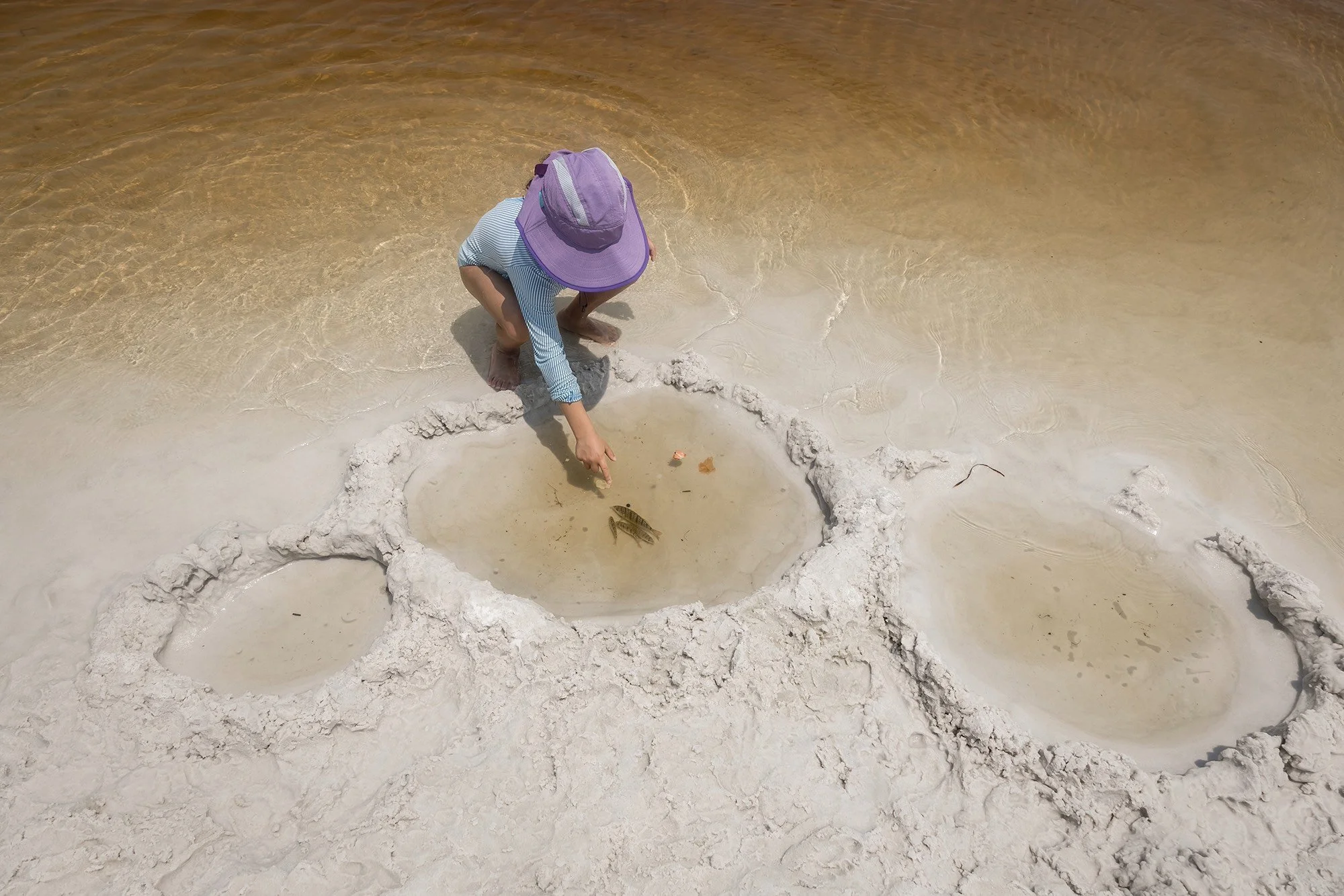 Unposed family photography at Grayton Beach State Park coastal dune lakes showing a child wearing a sun hat playing in the shallow water and sand.