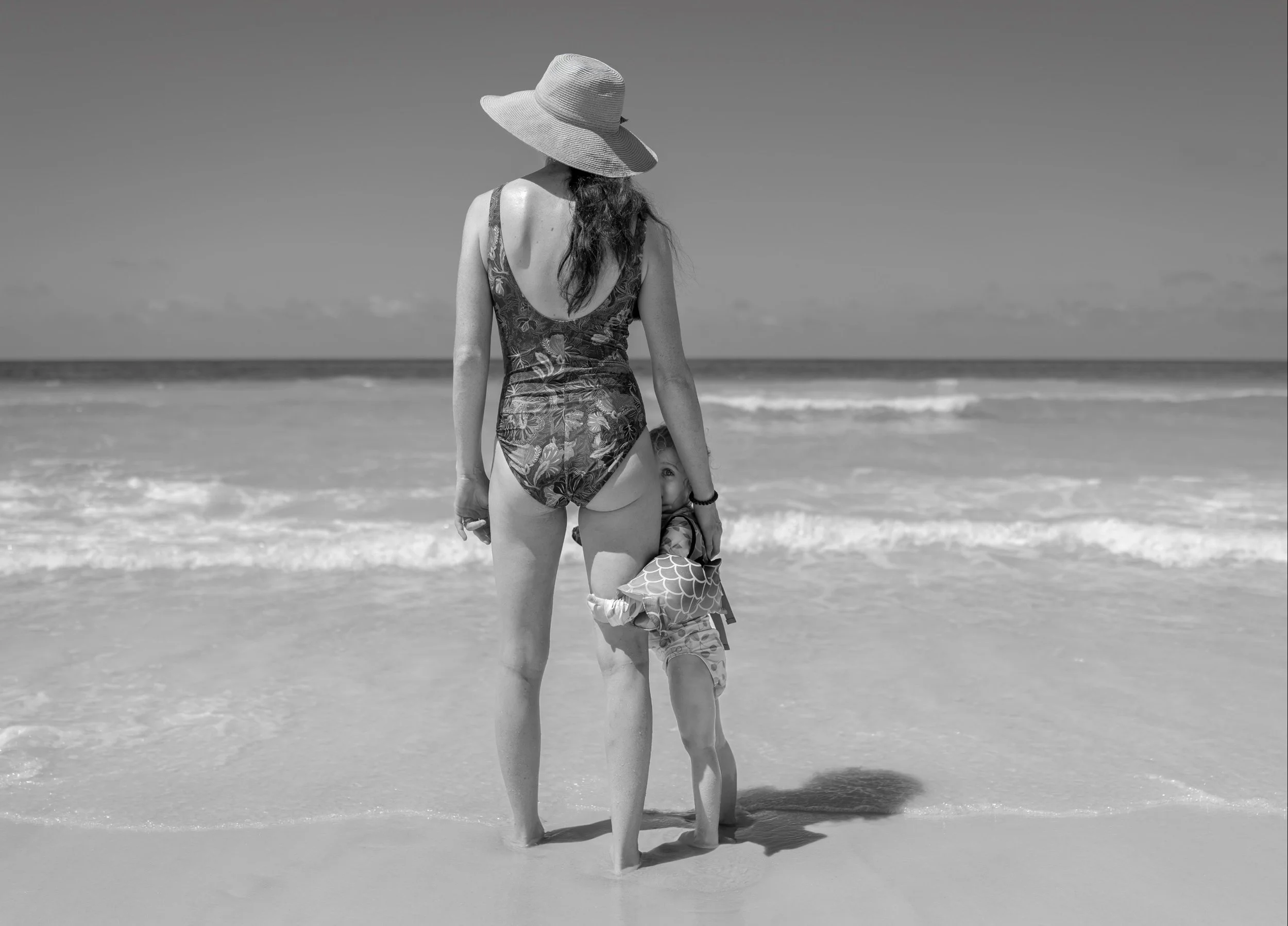 Toddler peeking out from behind mother's leg  on the beach during family photography session on 30A