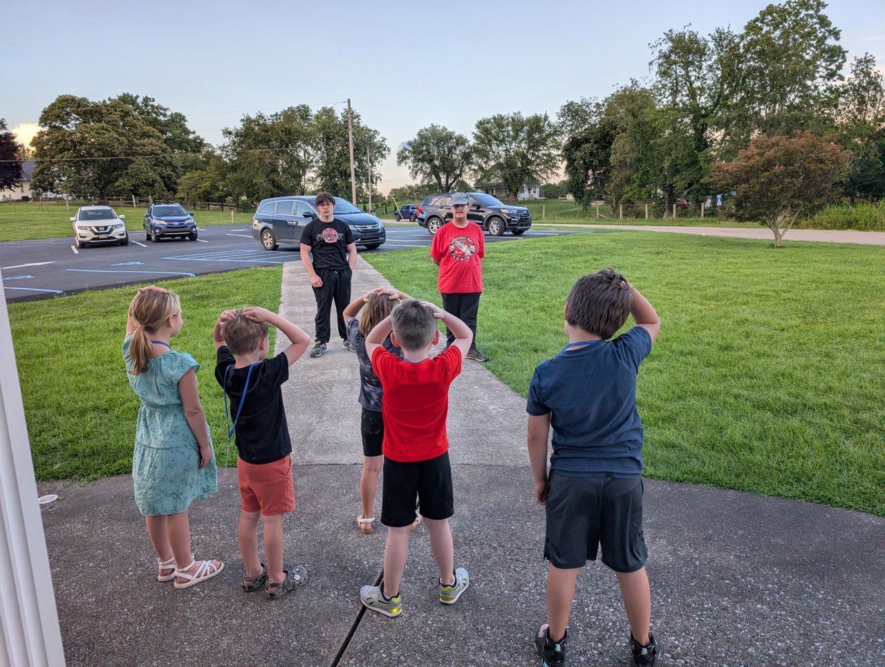Group of children standing outside on a sidewalk, saluting at two young men standing in front of them, with a parking lot and trees in the background.