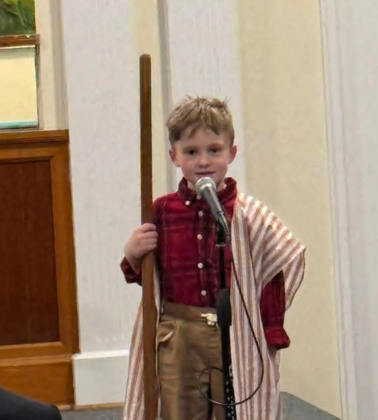 A young boy dressed in a plaid shirt, khaki pants, and a striped cape, standing in front of a microphone, holding a wooden staff, appearing to perform or speak at an indoor event.