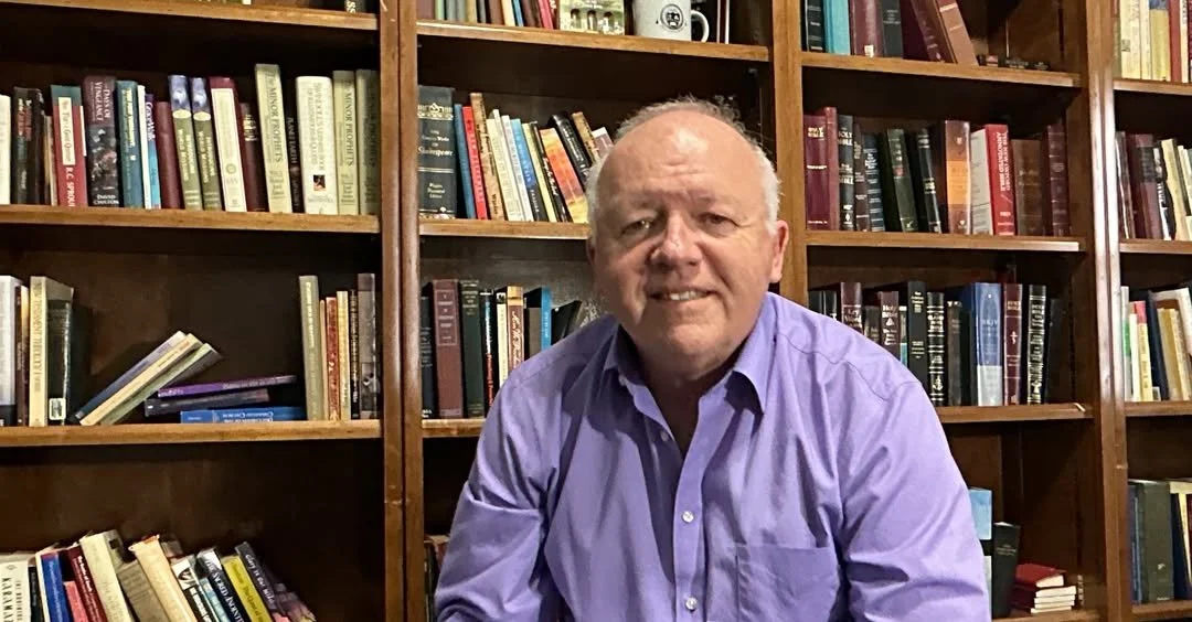 A middle-aged man with a receding hairline, wearing a purple button-up shirt, sitting in front of wooden bookshelves filled with various books.