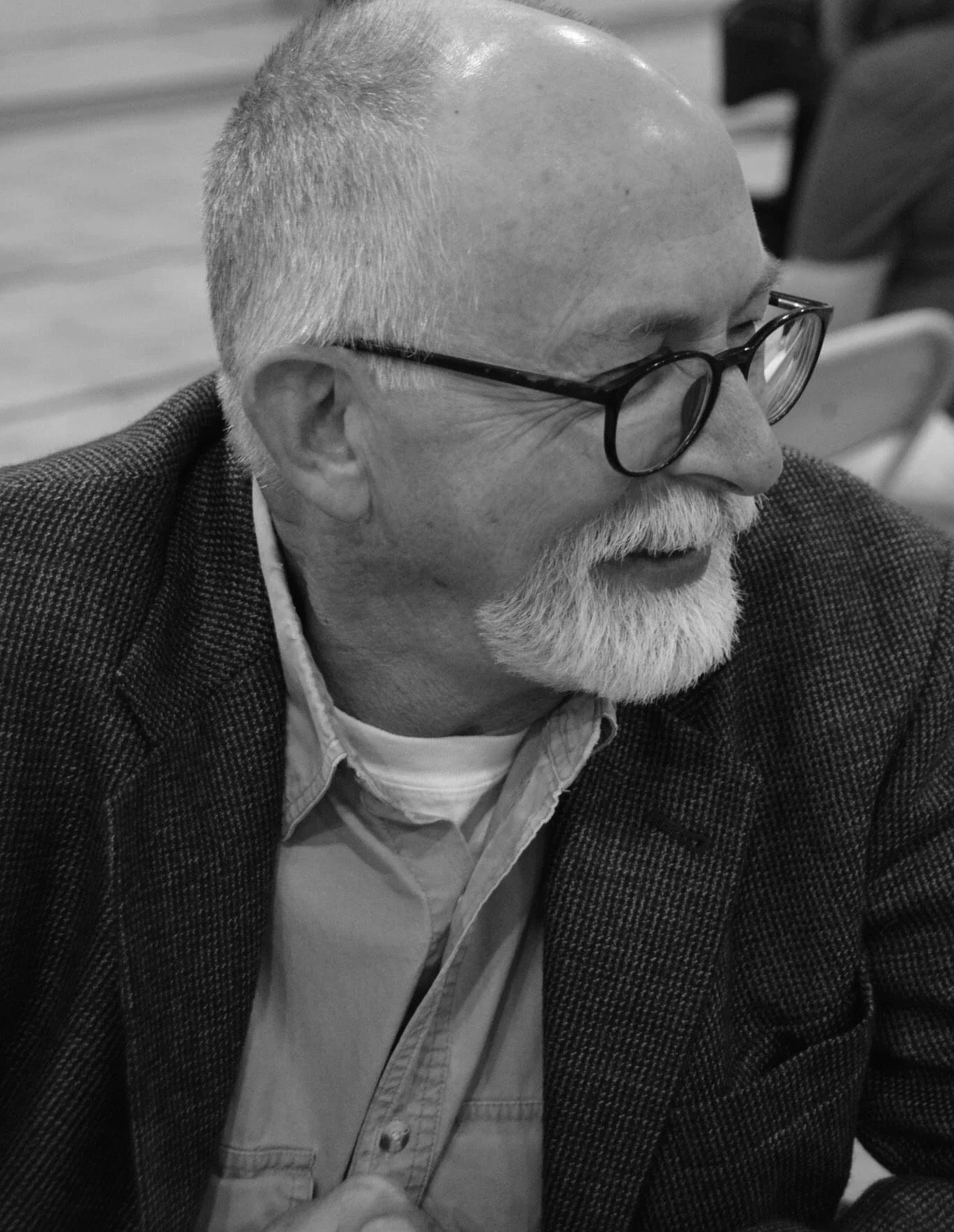 A black and white photo of an elderly man with glasses, a beard, and a shaved head, wearing a checked blazer and button-up shirt, sitting and looking to his right.