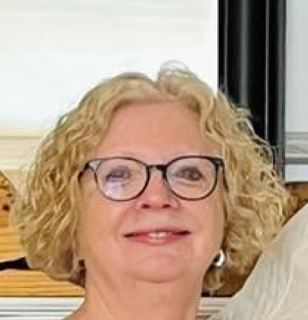 A woman with curly blonde hair and glasses smiling indoors.