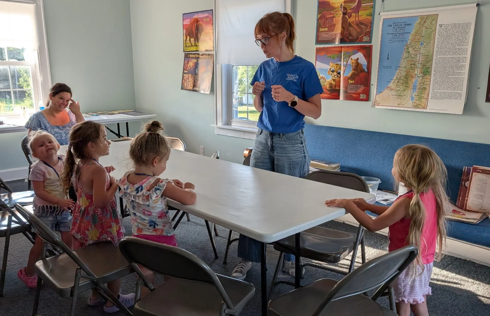 A woman in a blue shirt standing at a table with four young girls sitting and one girl standing, engaging in an activity in a room with educational posters on the wall.