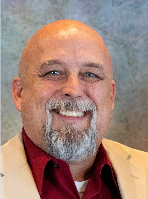 A smiling middle-aged man with a bald head, gray beard, and blue eyes, wearing a beige blazer over a red shirt, against a neutral background.