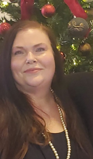 A woman with long brown hair smiling in front of a decorated Christmas tree