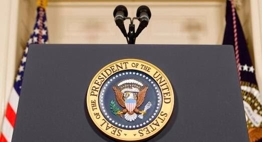 Photo of a podium with two microphones and the Presidential Seal on the front, two flags behind