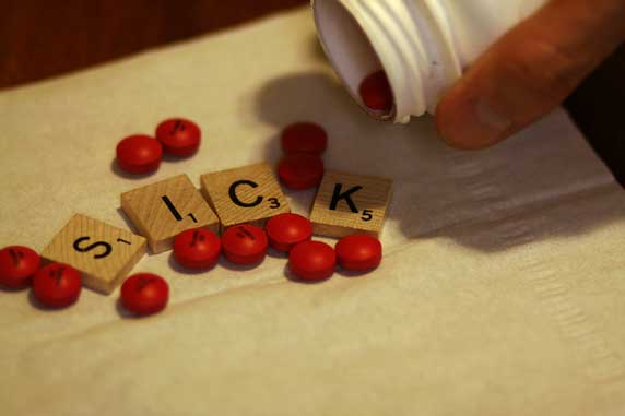 Photo of red pills being spilled onto a table, along with Scrabble tiles spelling "SICK"