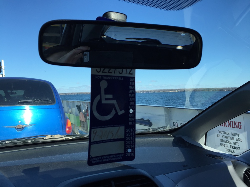 Photo description: View out the front window of a car on a ferry, from inside, showing the car parked in front, the ferry’s sides, and a view of the lake and blue sky. In the middle is the rearview mirror with a blue and white accessible parking permit hanging from it