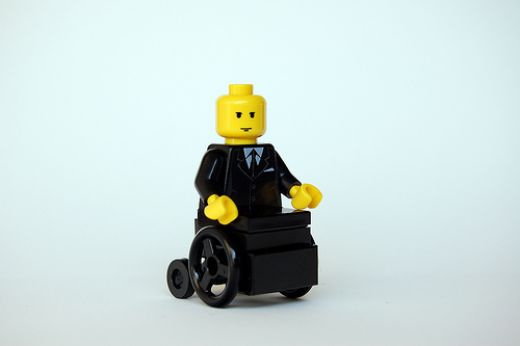 Lego figurine with yellow skin wearing a black suit and tie, sitting in a black Lego wheelchair