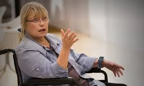 TV still image of a middle aged blonde woman sitting in a wheelchair, talking and gesturing