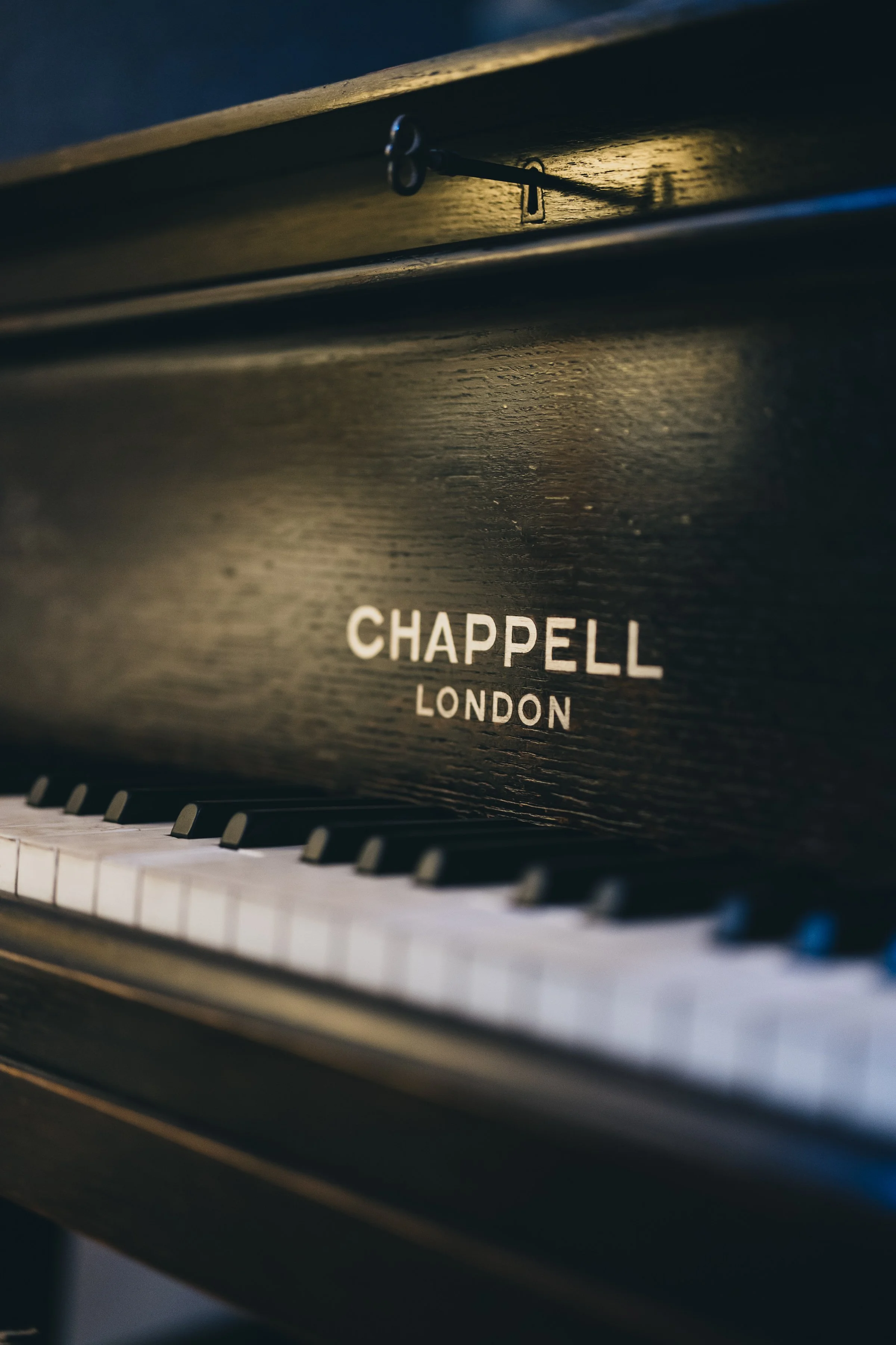 Close-up of a black upright piano with the words "Chappell London" on the front, showing a few white and black keys in the foreground.