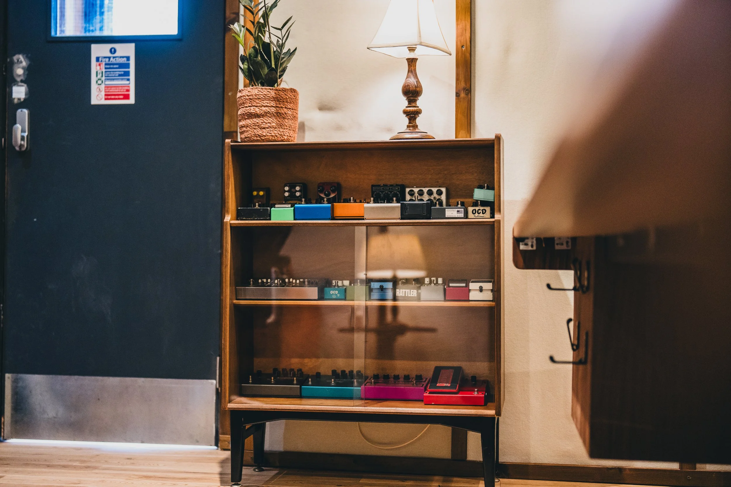 A wooden display shelf with multiple tiers holding various guitar effect pedals, positioned against a beige wall. A potted plant and a table lamp are on top of the shelf. Part of a kitchen counter and a blue door are visible nearby.