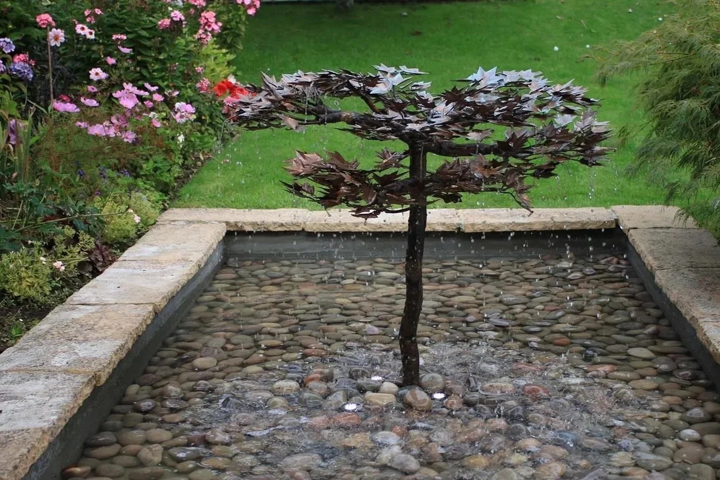 A small tree with dark leaves growing in a square fountain filled with round pebbles, surrounded by a garden with pink and purple flowers and green grass.