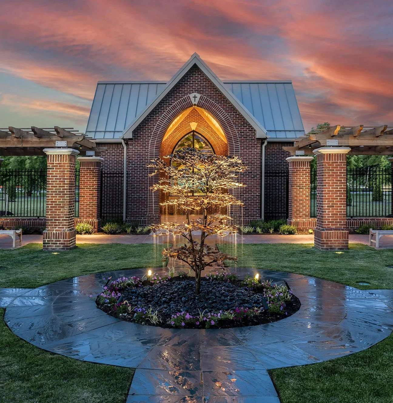 A small brick church with a pointed roof and a cross at the top, illuminated at sunset. In front, a landscaped garden features a tree with lights, a fountain, and flower beds on a stone pathway.