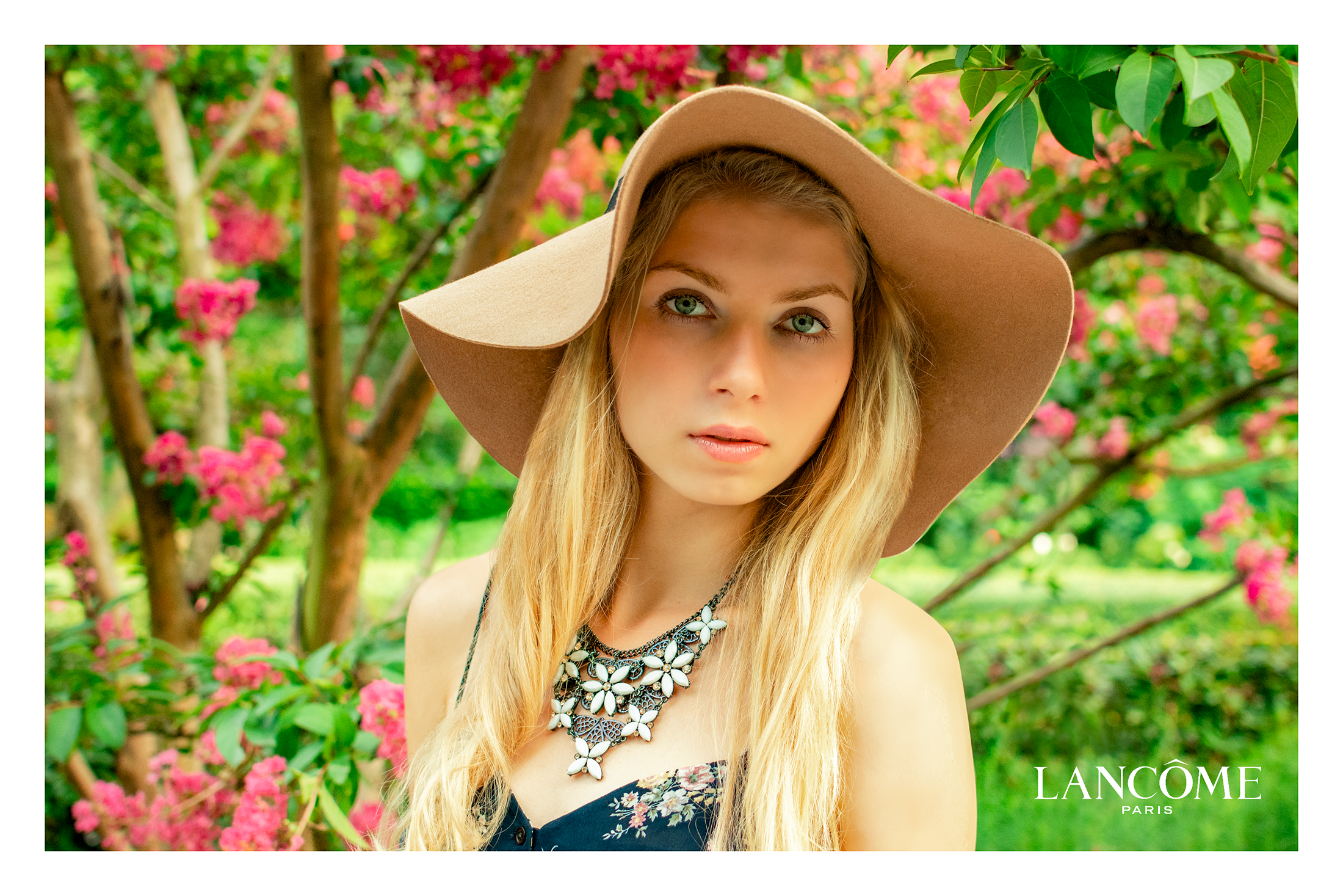 A young woman with long blonde hair wearing a beige floppy hat, a floral dress, and a large statement necklace, standing outdoors surrounded by pink flowering bushes.