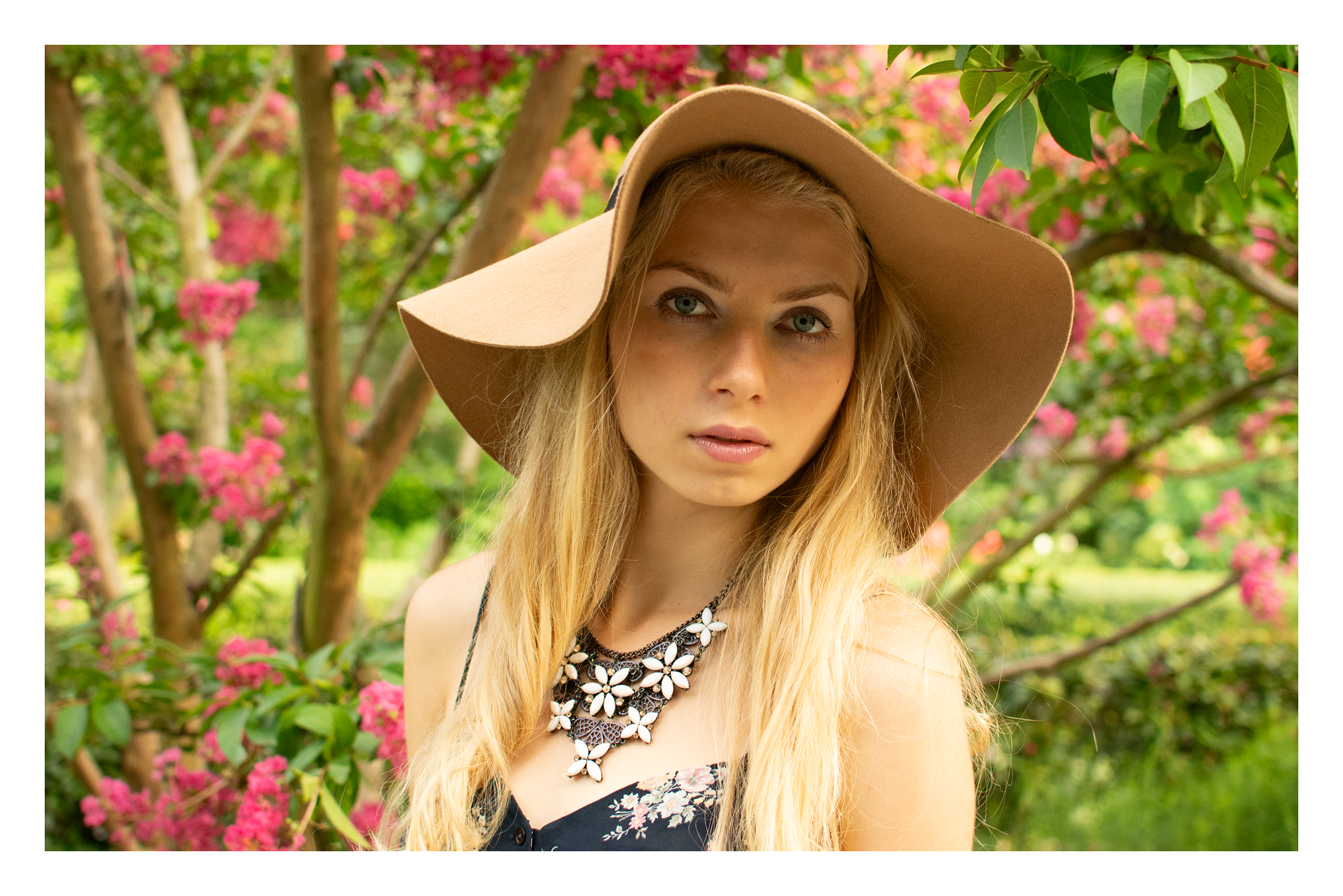 Young woman with long blonde hair wearing a large beige sun hat and a black floral top, standing outdoors with green foliage and pink flowers in the background.