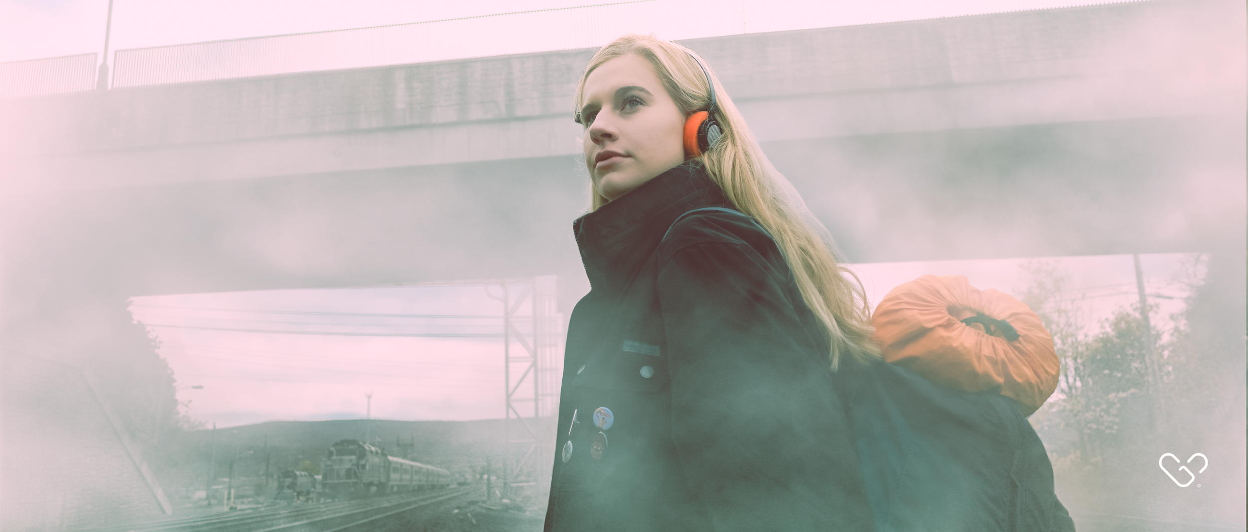 A young woman with blonde hair wearing a black jacket and red headphones, carrying an orange rolled-up sleeping bag on her back, standing near train tracks under a bridge on a foggy day.