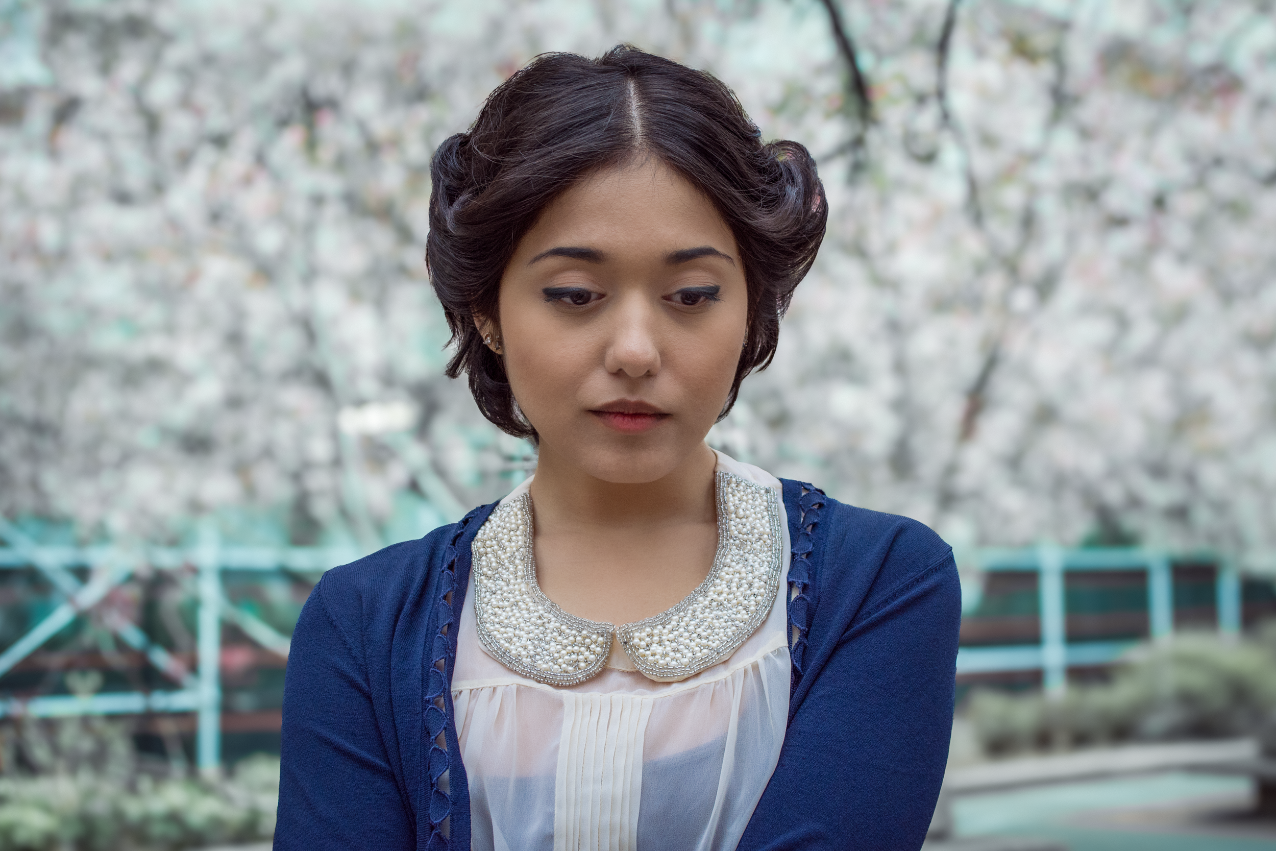 A woman with short dark hair wearing a cream top with an embellished collar and a dark blue cardigan standing outdoors with cherry blossom trees in the background.