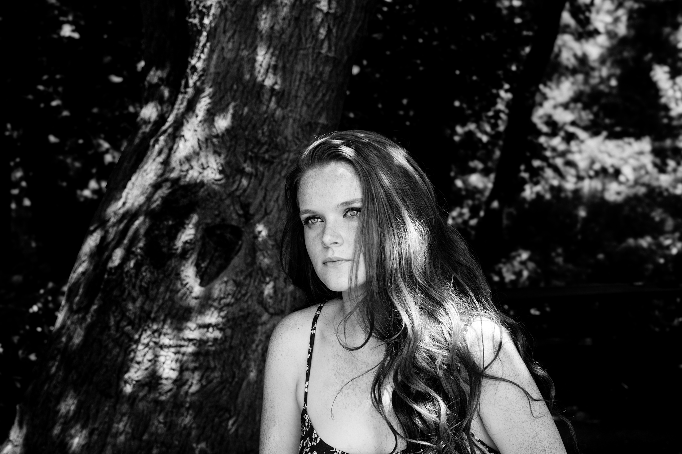 A young woman with long, wavy hair standing next to a large tree with textured bark in a shadowy outdoor setting.