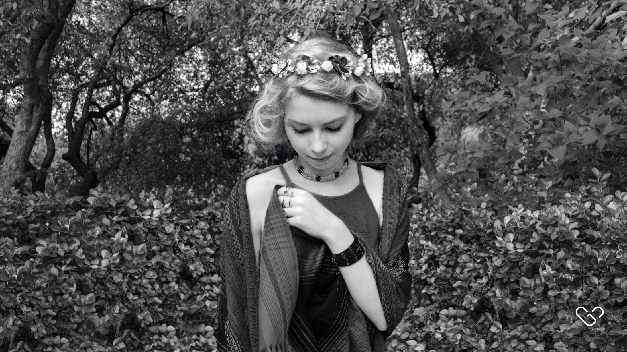 A young woman with short wavy hair wearing a flower crown and layered jewelry, looking down while standing among bushes and trees in a forested area.