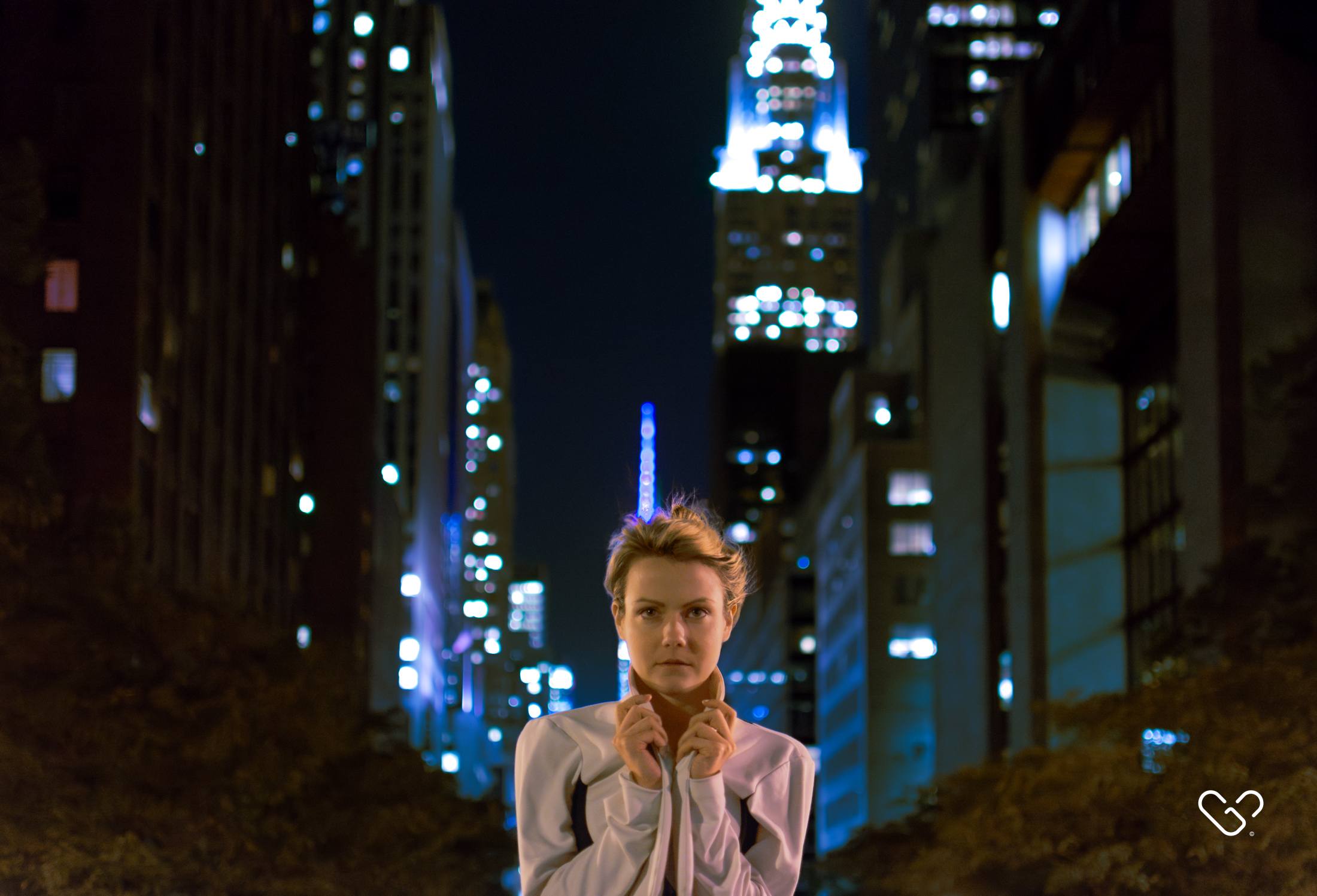 A woman standing in a city at night, with tall lit-up buildings in the background.
