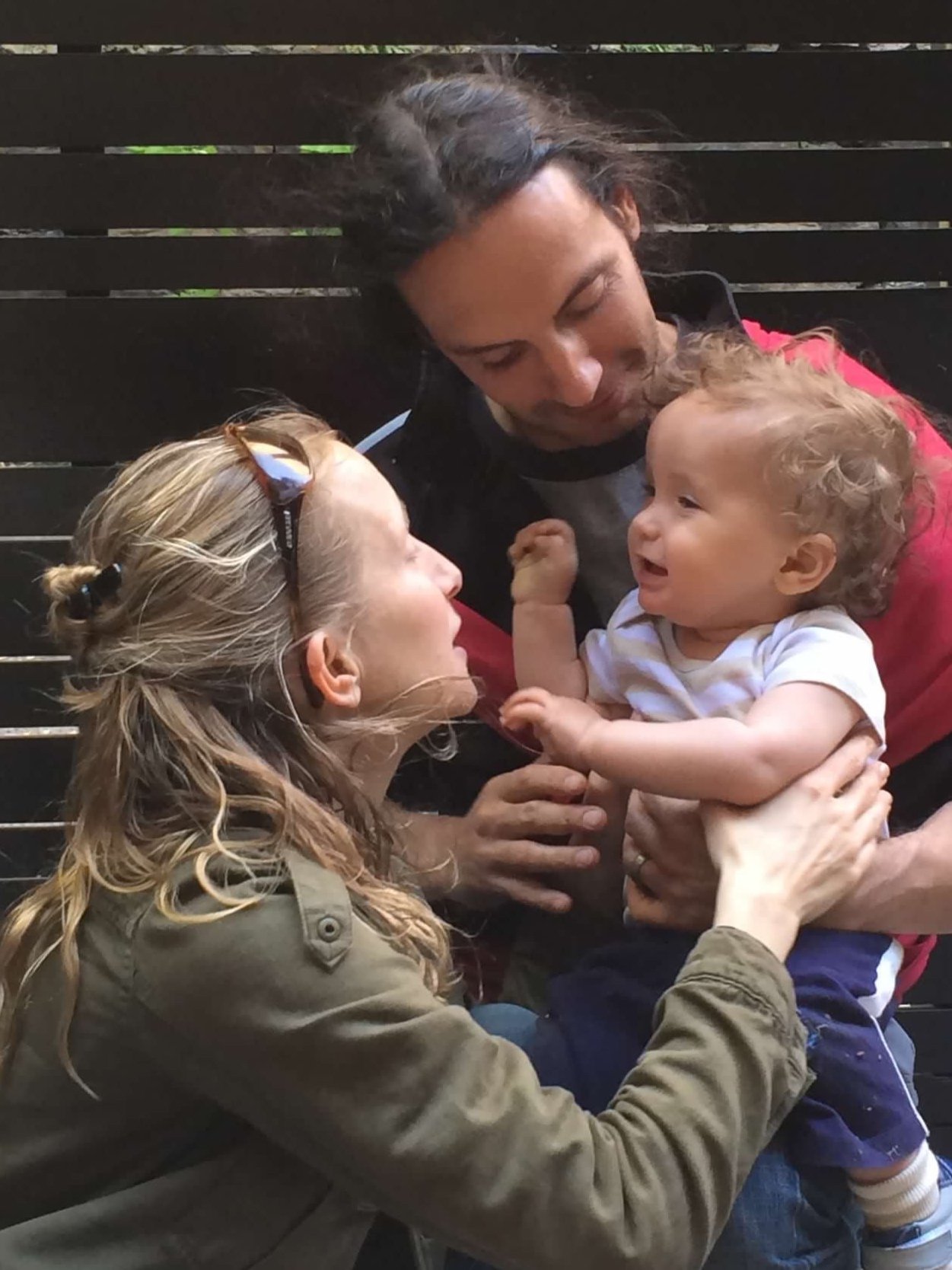 A mother, father and little boy. The mother is kneeling on the ground and the toddler is in his father's lap, reaching for her face. The mother is turned towards the child. She has long blond hair and sunglasses. The father is looking down at the boy