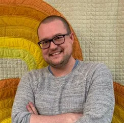 A smiling man with glasses and a beard, wearing a gray sweater, standing in front of a colorful rainbow fabric background.