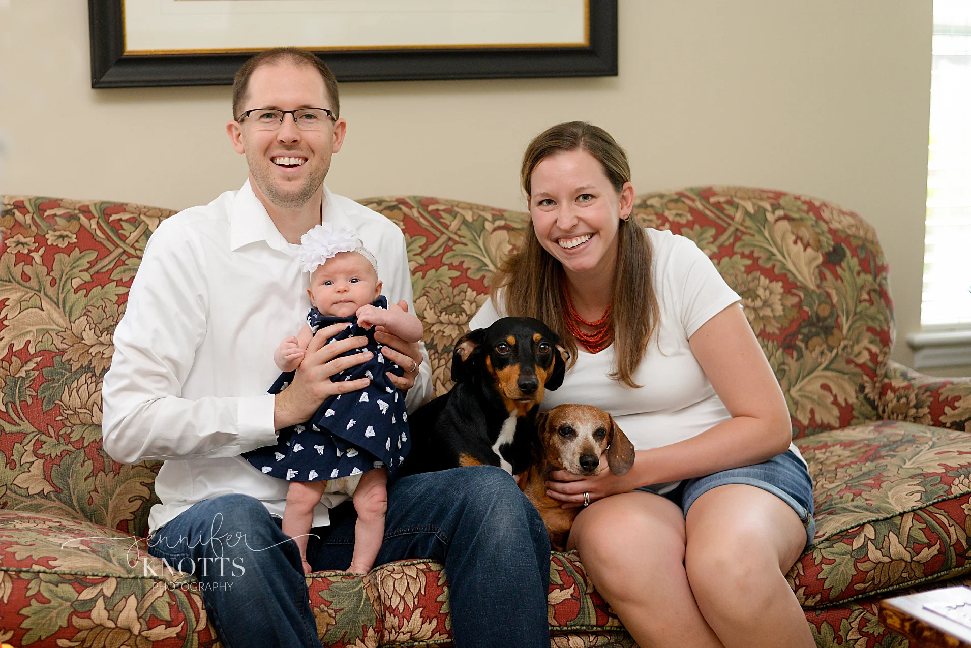 parents pose for photo with newborn daughter and two dogs on couch