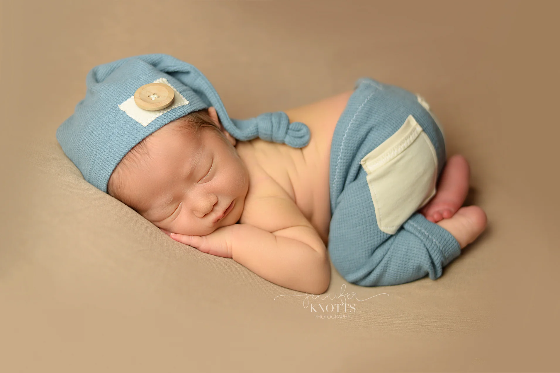 baby boy sleeps on tan blanket wearing blue outfit and hat