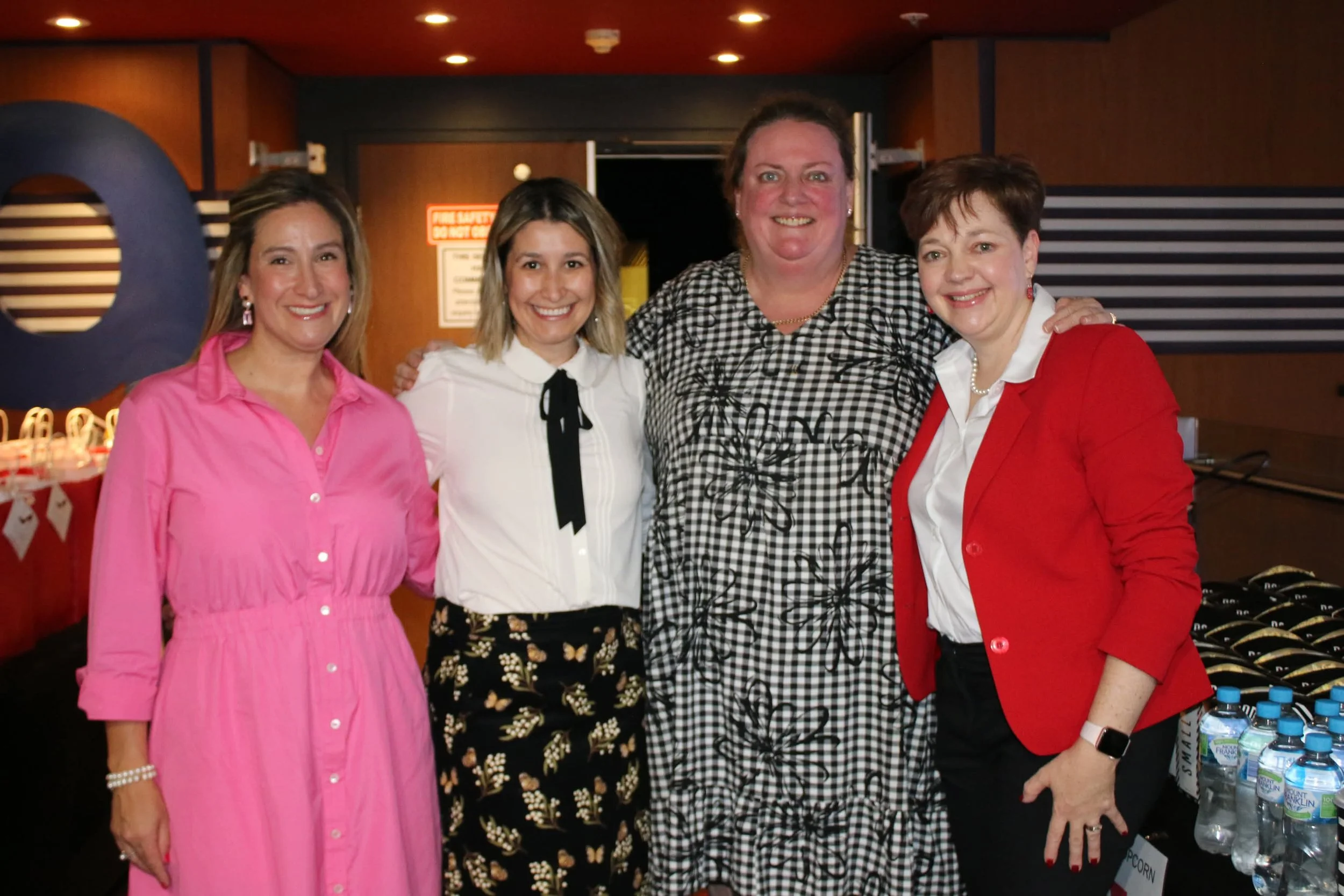 Four women standing together in an indoor space, smiling for the camera. One woman is wearing a pink dress, another in a white blouse with a black ribbon, one in a black and white checkered dress, and the last in a red blazer with white blouse. There are water bottles on a table to the right and a background with wooden walls and a fire safety sign.