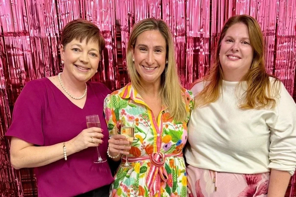 Three women smiling and holding champagne glasses, standing in front of a shiny pink backdrop.